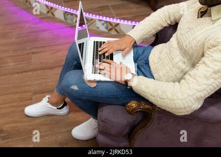 High angle of unrecognizable young Hispanic male in stylish outfit sitting on comfortable armchair working on laptop at home Stock Photo