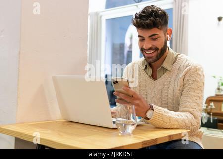 Smiling young Hispanic male with beard and dark hair sitting at home and browsing mobile phone Stock Photo