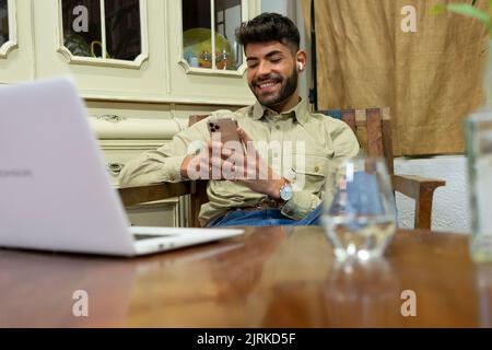 Smiling young Hispanic male with beard and dark hair sitting at home and browsing mobile phone Stock Photo