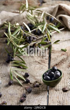 Small bowl of black pitted olives placed near green tree branches on shabby timber table in rustic kitchen Stock Photo