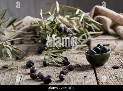 Small bowl of black pitted olives placed near green tree branches on shabby timber table in rustic kitchen Stock Photo
