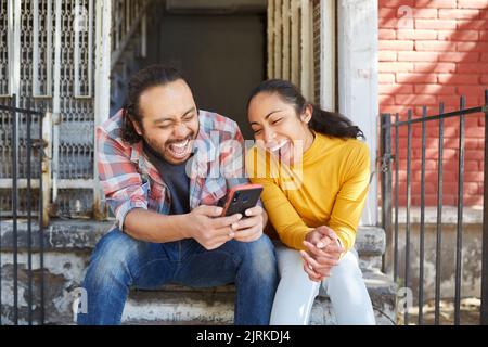 Young happy unshaven Mexican man with cellphone interacting with laughing female beloved while sitting on staircase in sunlight Stock Photo