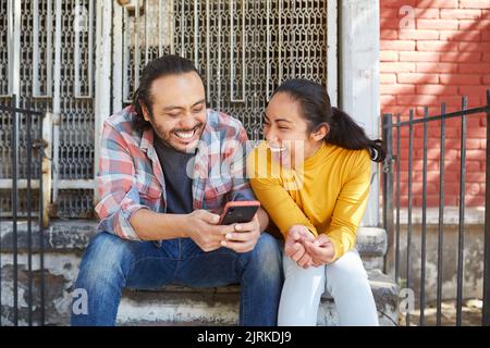 Young happy unshaven Mexican man with cellphone interacting with laughing female beloved while sitting on staircase in sunlight Stock Photo