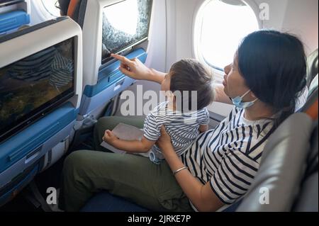 Infant traveling in airplane sitting on its mother lap using in flight entertainment screen. One year old baby boy flying in airplane and trying to en Stock Photo