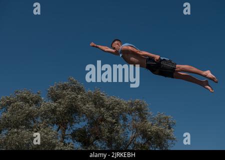 Young teen boy wearing a towel as a superhero scarf flying and diving in river. Clear blue sky and trees in distance as a natural background. Stock Photo