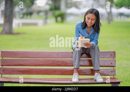 Full body of serious female in formal clothes sitting on wooden bench in park and writing in notebook Stock Photo