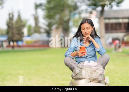 Weary female sitting with crossed legs and leaning on hand while browsing cellphone with frowning face in park Stock Photo