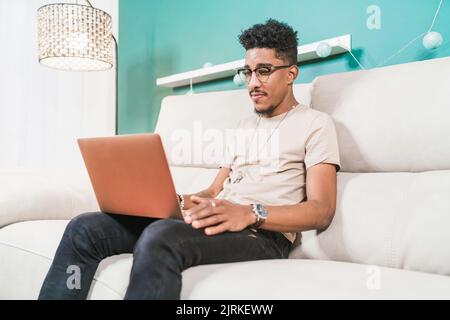 Young African American male remote worker sitting on couch and browsing netbook while working online Stock Photo