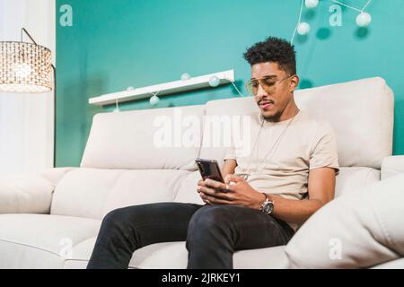 Young African American male browsing mobile phone while sitting on couch in modern apartment Stock Photo