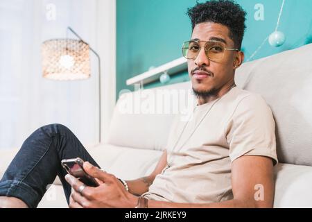 Young African American male browsing mobile phone while sitting on couch in modern apartment Stock Photo