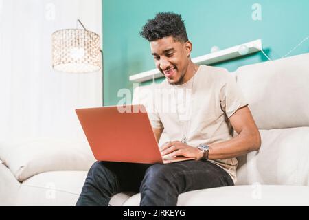 Young African American male remote worker sitting on couch and browsing netbook while working online Stock Photo