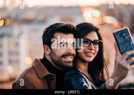 Content ethnic man with piercing near girlfriend taking self portrait on cellphone in town in twilight Stock Photo