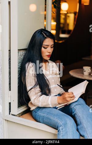 Young ethnic female in casual wear taking notes in diary while sitting on cafeteria windowsill Stock Photo