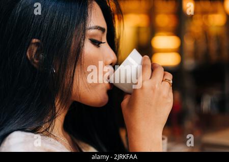 Side view of crop ethnic female with makeup drinking hot beverage from cup in coffee house Stock Photo