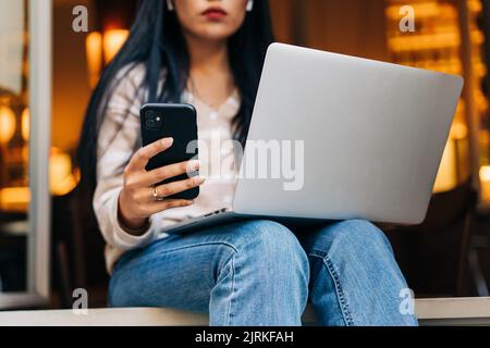 Low angle of young Asian female chatting on cellphone while sitting with netbook in cafeteria Stock Photo