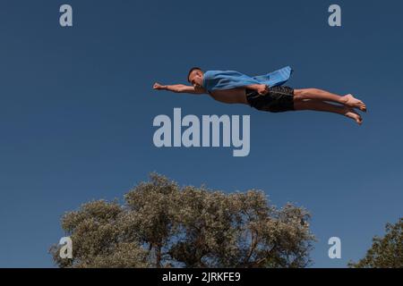 Young teen boy wearing a towel as a superhero scarf flying and diving in river. Clear blue sky and trees in distance as a natural background. Stock Photo