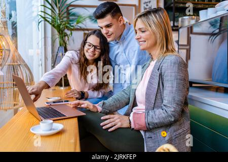 Content female employee pointing at netbook near male colleague and business trainer while talking at table in cafeteria Stock Photo