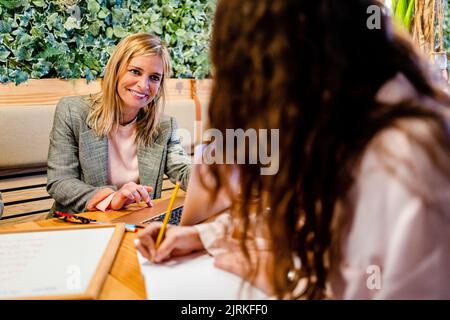 Adult content business trainer with laptop near crop anonymous female employee taking notes during coaching process Stock Photo