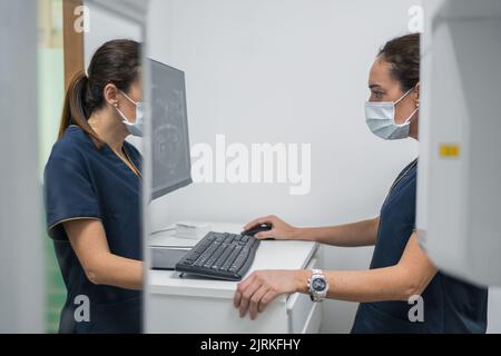 Side view of woman in medical uniform looking at teeth X ray on computer monitor while working in lab of contemporary dental clinic Stock Photo