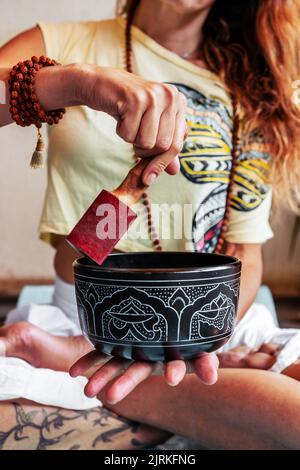 From above of crop female with traditional beads sitting in lotus pose and playing on Tibetan singing bowl during meditation and yoga practice Stock Photo