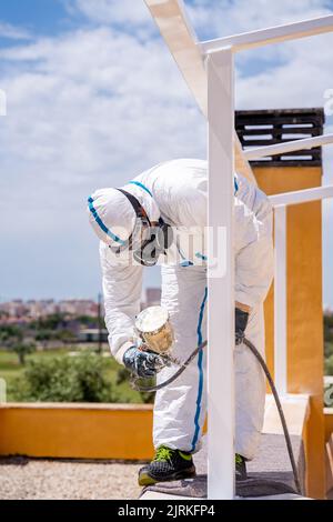 Anonymous employee in protective uniform and mask using air brush to paint frame structure against cloudy sky on sunny day Stock Photo