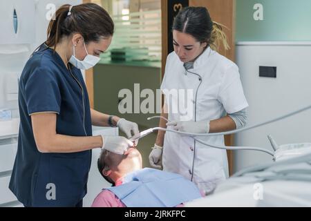 Woman in medical uniform using professional tool on teeth of adult male with nurse assistant working in contemporary dental clinic Stock Photo