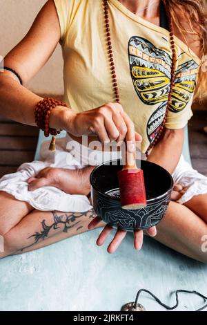 From above of crop female with traditional beads sitting in lotus pose and playing on Tibetan singing bowl during meditation and yoga practice Stock Photo