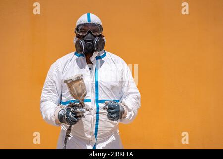 Male worker in white uniform and respirator holding air brush and looking at camera outside house during work Stock Photo