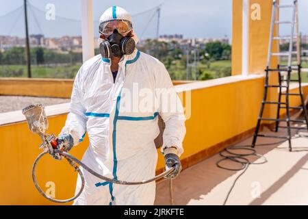 Man in white uniform and respirator carrying dirty air brush and looking at camera while working on building renovation site Stock Photo