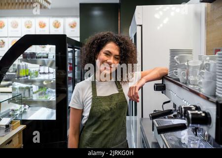 Delighted ethnic female barista in apron and with curly hair leaning on coffeemaker while standing in cafe and looking at camera Stock Photo