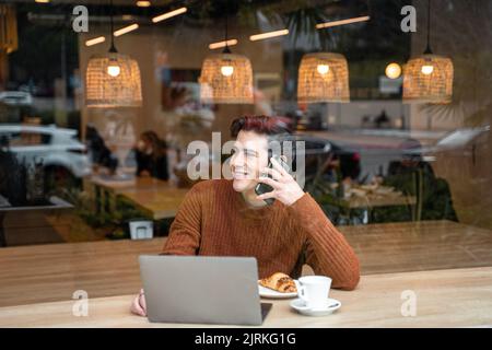 Through window of happy young male student sitting at table with laptop and cup of coffee with croissant and talking on mobile phone while spending ti Stock Photo