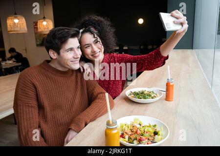 Cheerful young multiracial couple sitting at counter with healthy vegetarian salad and drinks and taking selfie on smartphone while having breakfast t Stock Photo