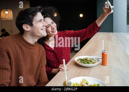 Cheerful young multiracial couple sitting at counter with healthy vegetarian salad and drinks and taking selfie on smartphone while having breakfast t Stock Photo