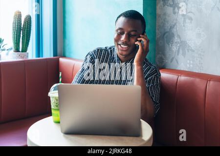 Content African American male entrepreneur speaking on cellphone against table with netbook and smoothie in restaurant Stock Photo