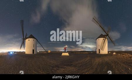 Back view of unrecognizable man standing with torch up on old traditional windmills located in countryside field on starry night Stock Photo