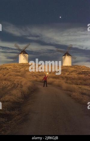 Back view of unrecognizable man standing with torch up on old traditional windmills located in countryside field on starry night Stock Photo