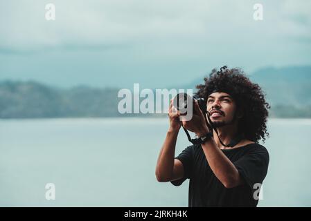 Ethnic male with Afro hairstyle taking photo of ocean on digital camera on urban embankment Stock Photo