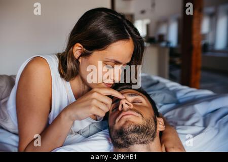Calm young woman with long hair gently touching nose of sleeping man while lying together on bed at home in daylight Stock Photo