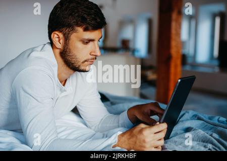 Side view of young bearded guy touching screen of tablet while lying on bed in light modern apartment Stock Photo