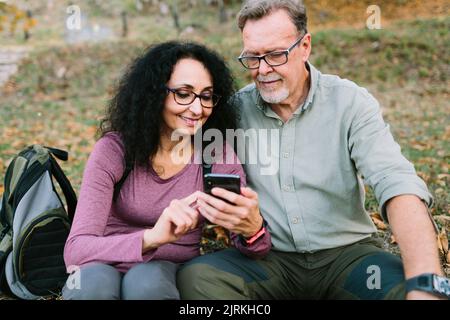 Content senior couple in casual clothes and eyeglasses browsing modern smartphone while sitting together on grassy ground in autumn park Stock Photo