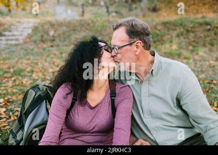 Cheerful mature couple in casual wear sitting and kissing on ground in autumn park Stock Photo