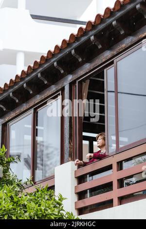 Low angle of elderly female looking away and thinking while peeking out from window of balcony at home Stock Photo