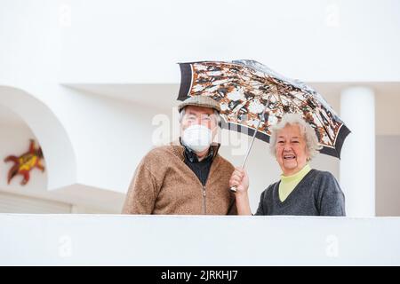 Cheerful elderly woman smiling and keeping umbrella over man in medical mask while standing on terrace of estate Stock Photo