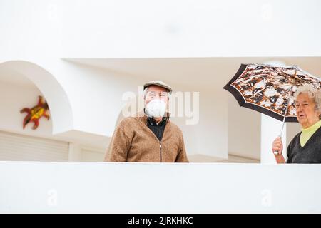 Elderly man with medical mask while standing on the terrace of the farm with his wife carrying an umbrella in quarantine Stock Photo