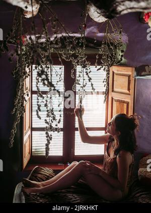 Side view from above of young woman in casual t shirt sitting on couch and touching soft plants hanging from ceiling with window on background Stock Photo