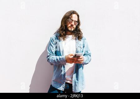 Bearded ethnic male in denim jacket and modern sunglasses surfing internet on cellphone in sunlight on white background Stock Photo