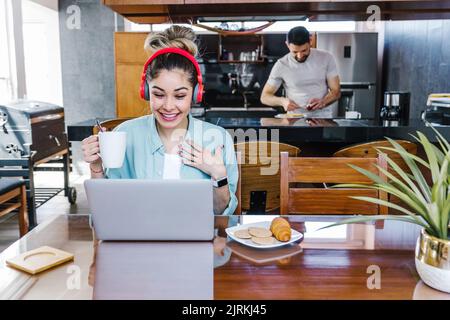 Cheerful Latin female in headphones watching funny movie on netbook and having breakfast at home on background of blurred boyfriend cooking in kitchen Stock Photo
