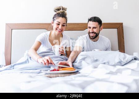 Low angle of happy ethnic couple chilling in bed and enjoying tasty breakfast in morning at home Stock Photo