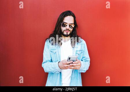 Bearded ethnic male in denim jacket and modern sunglasses surfing internet on cellphone in sunlight on red background Stock Photo