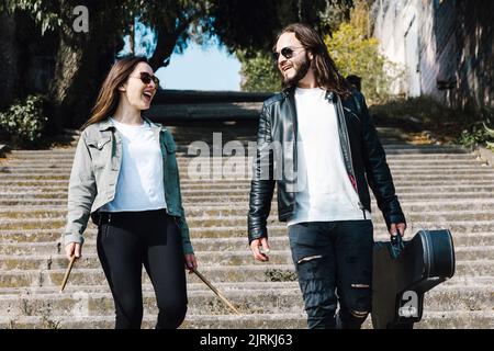 Latin American male musician holding acoustic guitar case while walking and looking at female beloved with drumsticks on urban staircase Stock Photo